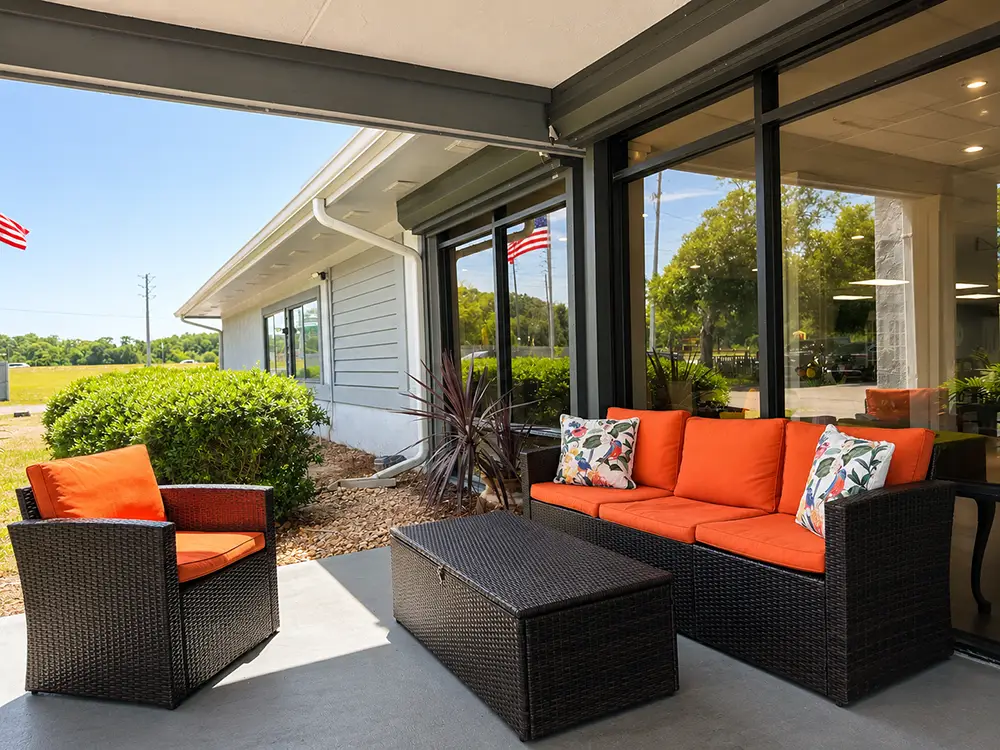 Wicker patio furniture with orange cushions and floral pillows sits on a covered porch outside a building with large windows on a sunny day.