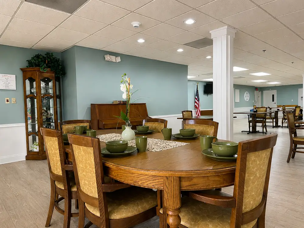A dining room with a round wooden table set for six, green dishes, a vase with flowers, wooden chairs, a piano, and an American flag in the background.