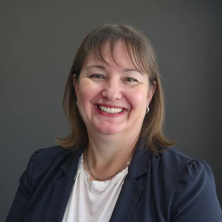 Woman with shoulder-length brown hair, wearing a navy blazer and white blouse, smiling in front of a plain gray background. Woman with shoulder-length brown hair, wearing a navy blazer and white blouse, smiling in front of a plain gray background.
