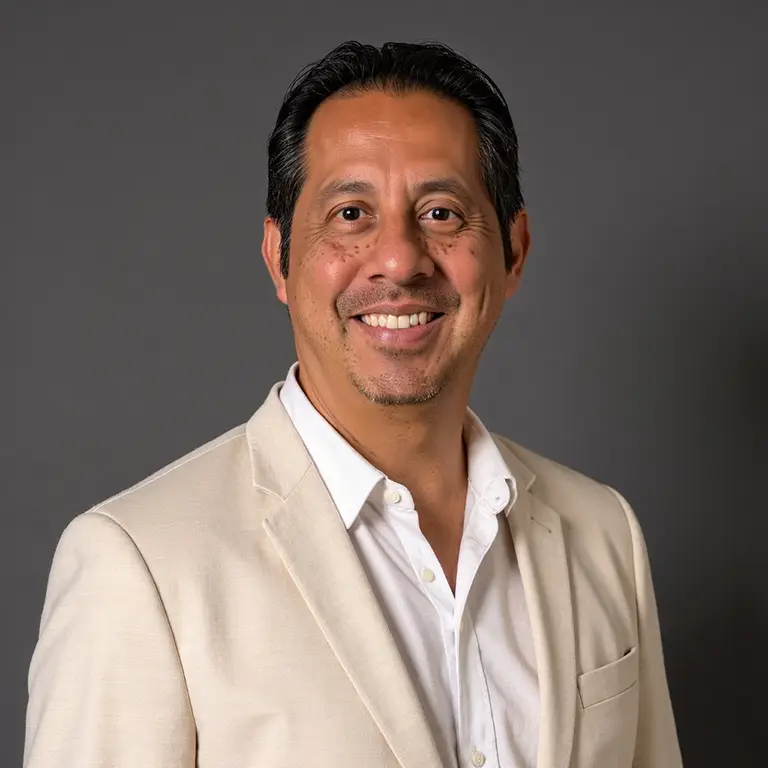 Man with short black hair, goatee, and beige suit jacket over a white shirt stands smiling in front of a plain gray background.