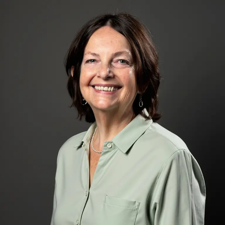 Woman with shoulder-length brown hair, wearing a light green button-up shirt, a necklace, and earrings, smiling against a plain dark background.