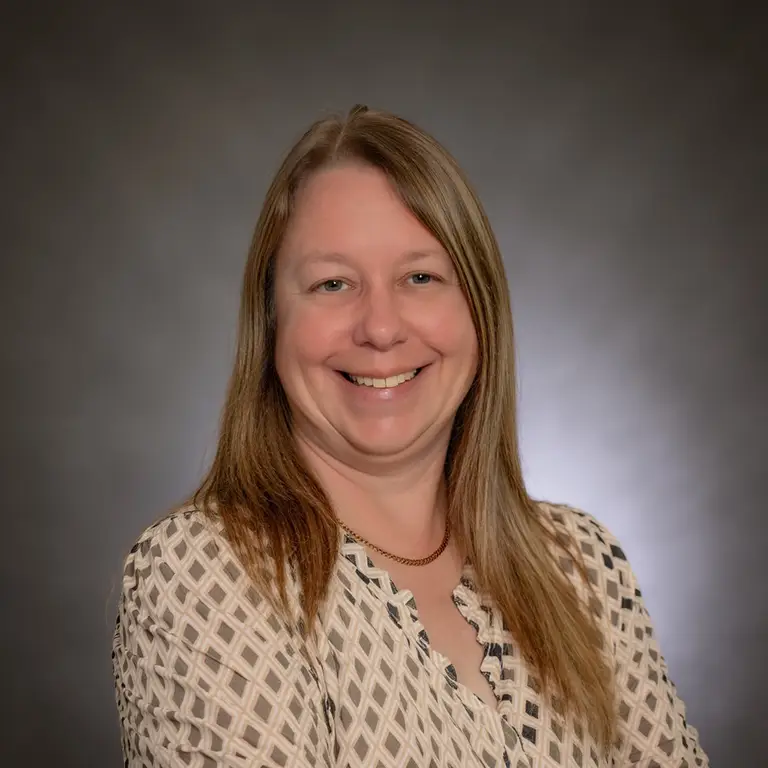 Woman with long light brown hair, wearing a patterned blouse and gold necklace, smiling in front of a neutral gray background.