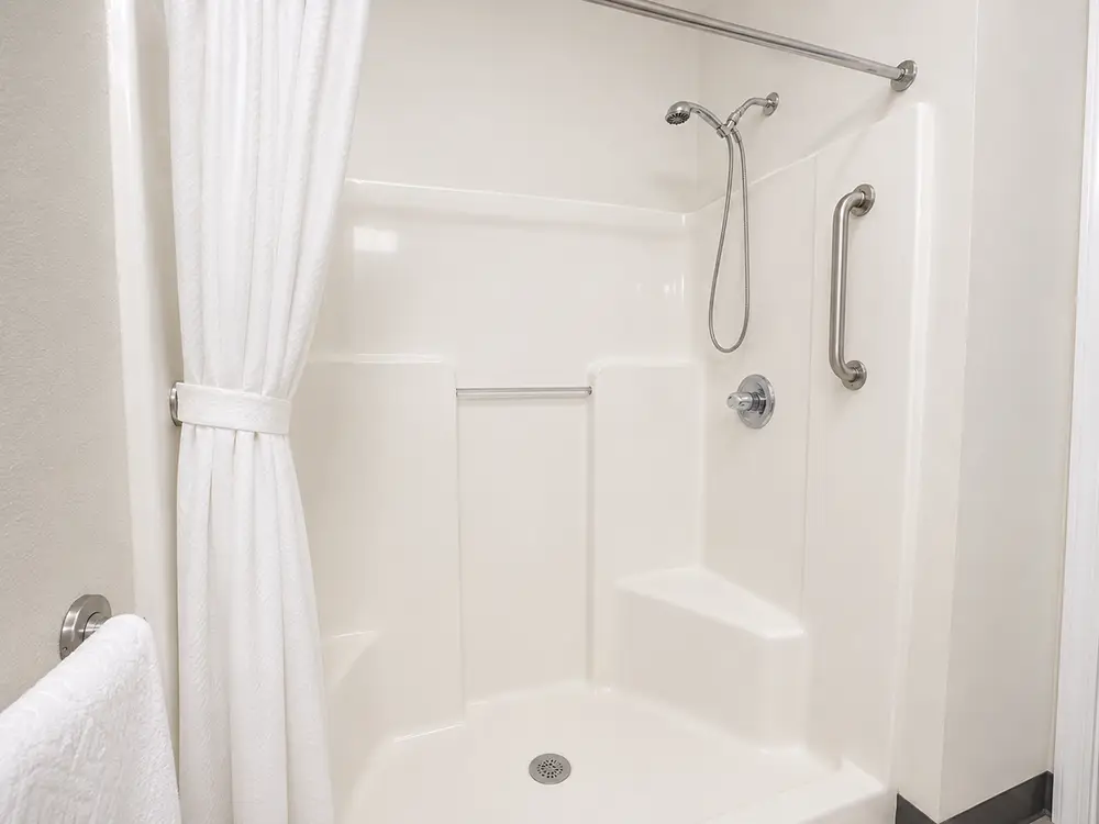 Accessible shower with grab bars, built-in seat, handheld showerhead, white walls, and a white shower curtain.